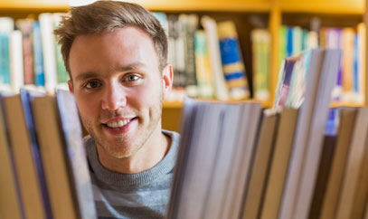 Ein junger Mann schaut zwischen Büchern durch ein Bücherregal.